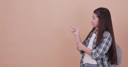 Cute school girl is carrying a school bag and pointing something. Isolated on brown background
