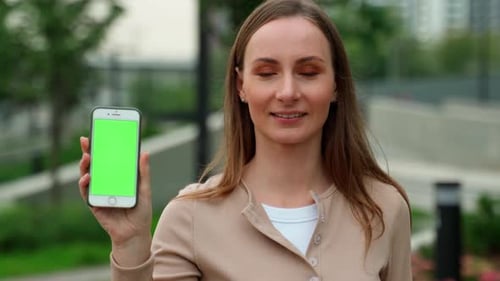Young Cheerful Woman Shows Off a Smartphone with a Green Screen While Standing Outside