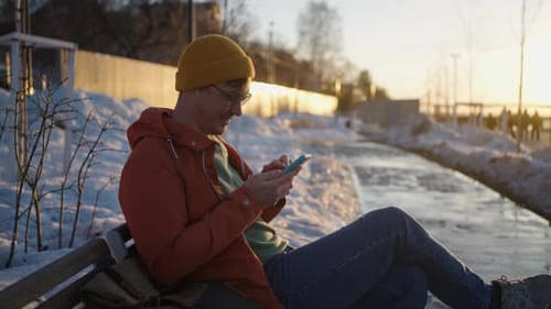 Young Man Relaxing in Warm Beanie and Glasses Browsing Smartphone While Sitting on Park Bench During