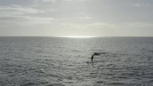 Silhouette stand up paddleboarder on flat grey ocean near rock hazard