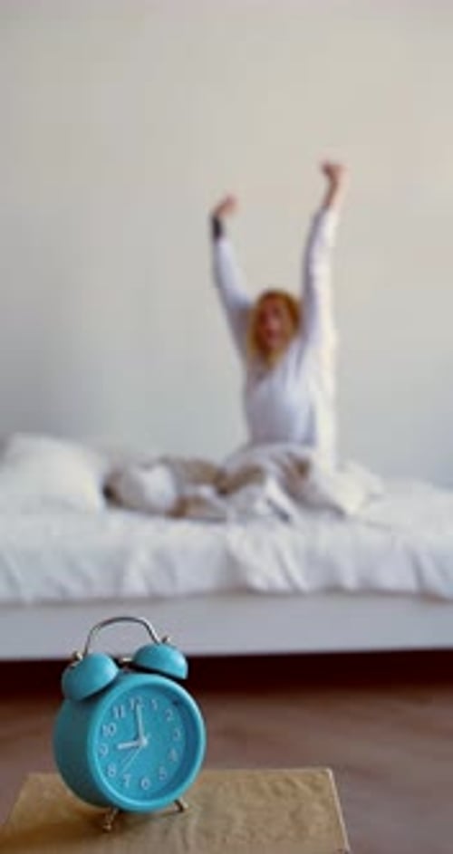 Vertical Video of an Alarm Clock on Foreground and a Blurred Young Blonde Woman Woke Up and