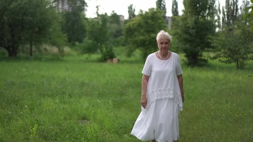 Slow Motion A Tired Elderly Upset Woman in a White Dress Leisurely Walks Along the Green Grass in a