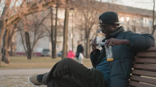 Man Sitting on Park Bench Eating Snack and Drinking Juice