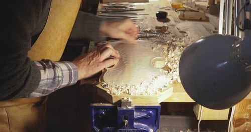 Luthier working on the top plate of a violin using a gouge to smooth in his workshop, top down view