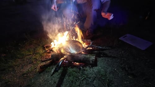 A man cooks a bream on a frying pan over a fire in Australia at night.