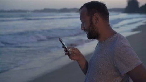 Young Man Texting on Smartphone Standing Near Sea in Evening Adult