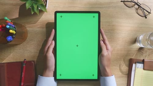 Top Down View Of Man's Hands Holding A Green Screen Tablet On A Wooden Desk