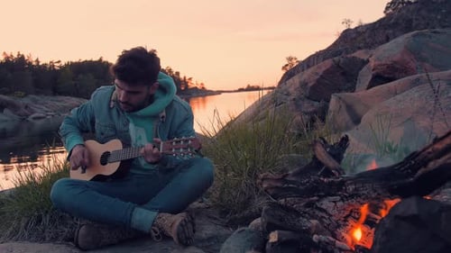 Jeune homme chantant avec une guitare acoustique devant un feu de joie et la mer sur une belle île isolée.