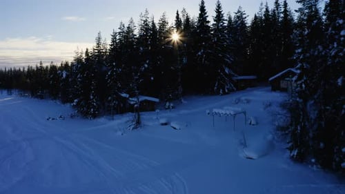 Aerial view rising over sunrise shining behind peaceful snow covered winter woodland cabins