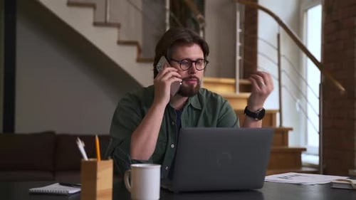 Young Man Talking on Phone in Front of Laptop and Sitting at Table in Home Office Spbas