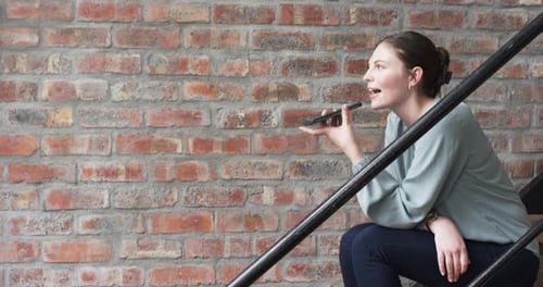 Businesswoman records voice message on stairs, green blouse, brick wall background.