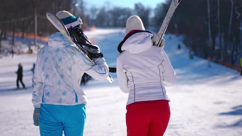 Back View Two Happy Confident Young Women with Ski and Snowboard Walking on Snowy Slope on Winter