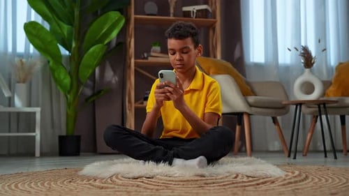 Teen Boy Uses Smartphone Sitting Cross-Legged on Rug