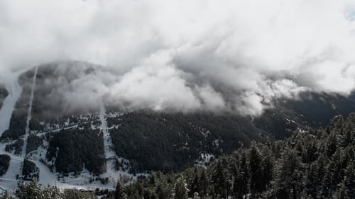 Aerial panorama between trees of a valley in cloudy alpine snowy mountain with clouds. Moody scene