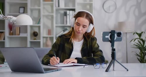 Young college girl student in headphones sitting table writing notes by looking into mobile phone