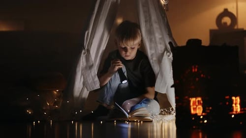 A Cute Blonde Boy Reads a Book By the Light of a Lantern Sitting at Home in a Hut in the Dark By the