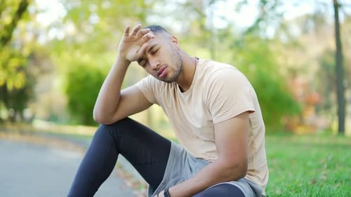 Close up of a young runner in a t-shirt overtired after a hard run sitting in an urban city park.