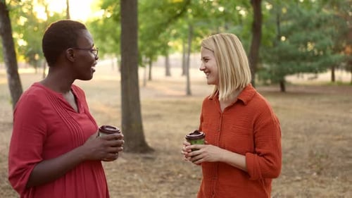 Two diverse women enjoying a conversation while holding coffee cups in a beautiful park