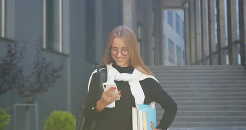 Smiling woman in glasses which holding books in hands and looking using smartphone near building