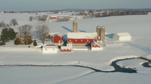 Aerial of red barn and farm buildings in winter snow. Stream winds through field. Overhead drone vie