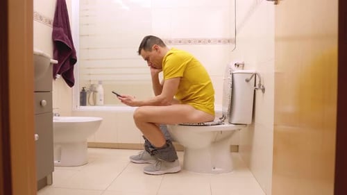 Man Sitting on Toilet Using Smartphone in Bathroom