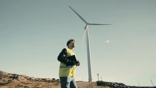 Eine Weitwinkelaufnahme eines jungen männlichen Ingenieurs, der auf eine Windturbine zugeht und dann Technologie auf einem T einsetzt