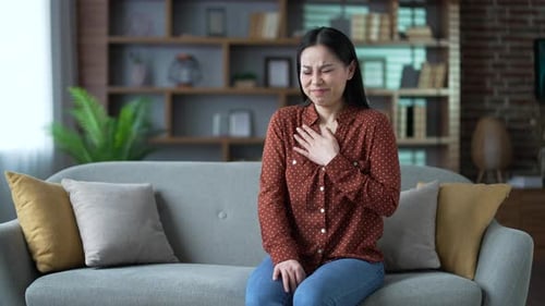 Asian woman has a heart attack while sitting on sofa in living room at home. Sick female suffers
