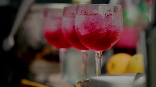 Three glasses of red cocktails with ice, lined up on a bar with fruits blurred in the background.