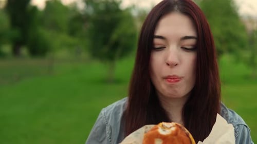 Portrait of Caucasian Woman Eating Cheeseburger in the Park Filmed Against Green Grass Background