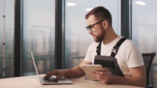 Portrait of technician in work uniform and glasses diagnoses laptop in modern office with large wind