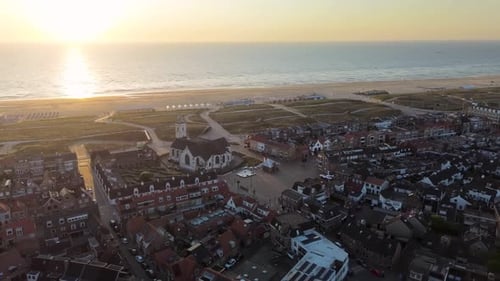 Aerial view of a coastal town at sunset with a white church, red-roofed houses, grassy dunes