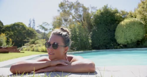 Relaxing by pool, woman wearing sunglasses and leaning on edge, enjoying sunny day, copy space