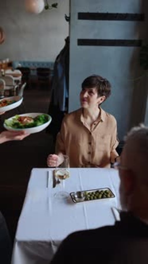 Pensive blonde waitress brings food for cheerful elderly couple in cafe.