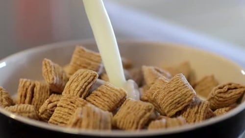 Close up view of milk pouring into a full bowl of tasty cereal while adding a silver spoon.