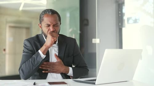 Man Coughing at Desk in Bright Office