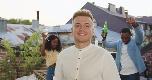 Portrait of smiling young man looking at camera near his positive dancing friends on terrace