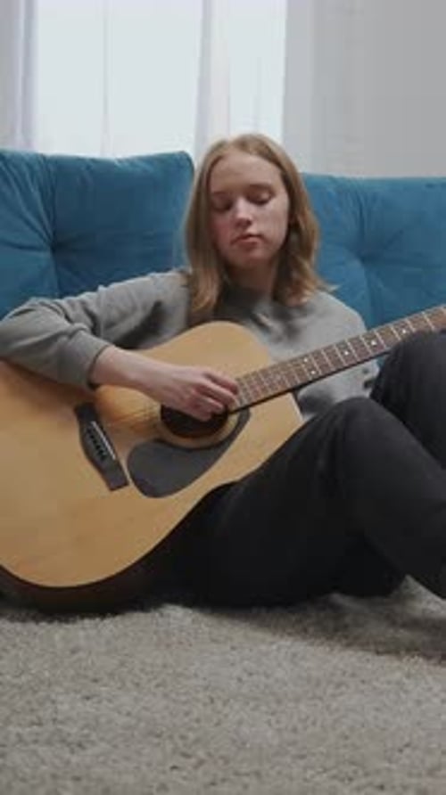 Teen Girl Sitting on Living Room Floor Playing Acoustic Guitar By Blue Sofa Practicing Relaxing