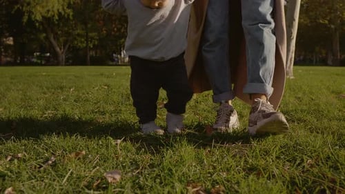 Mother supports her young son as he takes his first steps outside in the park