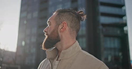 A Young Man with Stylish Trendy Hairstyle and Beard in Casual Clothes Stands on the Street in the