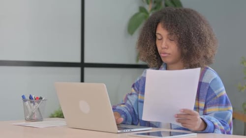 Young Woman Reviews Paperwork at Desk with Laptop