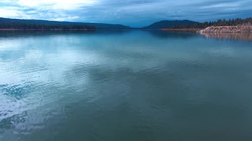 Smooth low altitude drone flyover across blue Lake with mountain silhouettes and calm reflections