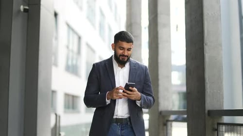 Happy smiling businessman in formal suit is using phone walking near an office building. Handsome