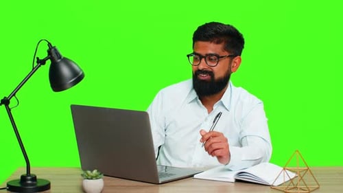Indian Man Writing Ideas in Notebook While Working on Laptop Focused Multitasking Planning Concept
