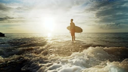 Surfer steht im Meer und hält Surfbrett bei Sonnenuntergang
