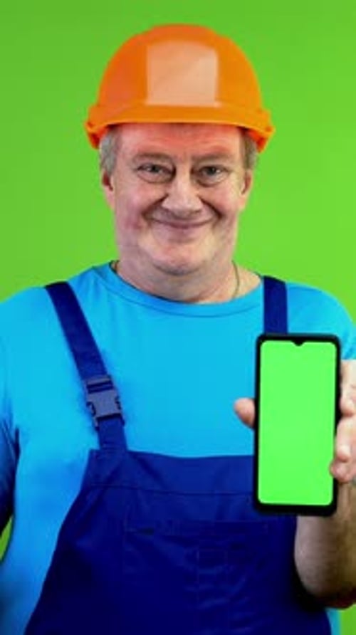 Senior Construction Worker in Overalls Pointing with Finger at Green Display and Smiling