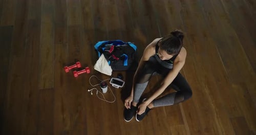 Top view of a fit woman seated on the floor of a gym fastens her shoes, with water bottle of an en