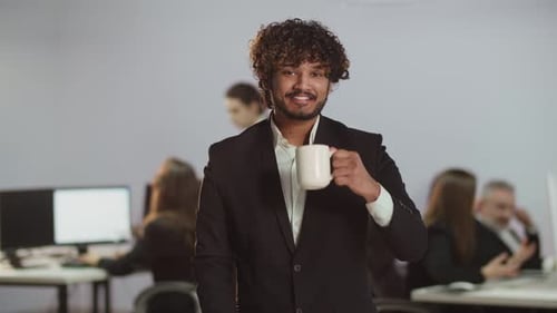 A Young Indian Man in a Business Suit is Drinking Coffee and Looking at the Camera View of a Smiling
