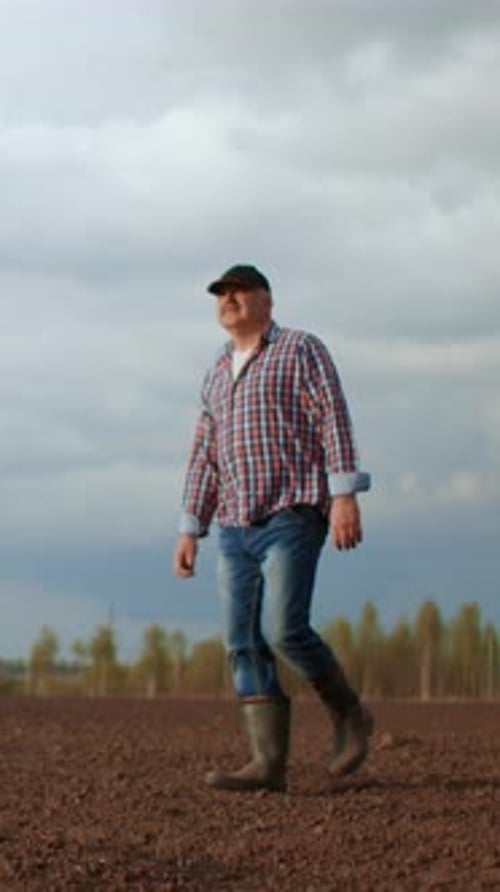Confident Farmer in Plaid Shirt and Rubber Boots Walking Across Freshly Tilled Agricultural Field