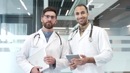 Portrait of two smiling doctors standing in modern hospital clinic looking at camera.