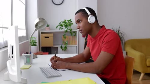 Young African Man Sitting at Desk Working on Laptop Taking Notes in Notebook